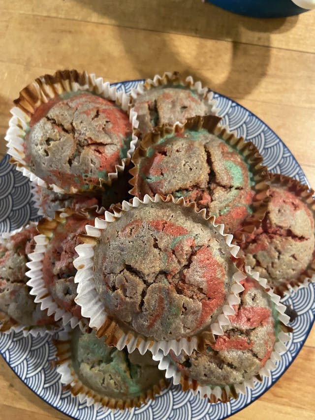 Alt text: A plate of freshly baked muffins with a swirled mix of colors, including shades of red and green, suggesting a possible berry or fruit flavor. They are presented in paper baking cups and sit on a decorative plate with a visible blue pattern.