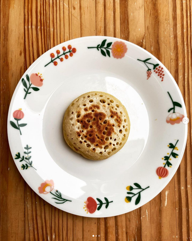 A single, golden-brown pancake with a pattern of small holes on the surface, centered on a white plate with floral and berry patterns, placed on a wooden table.